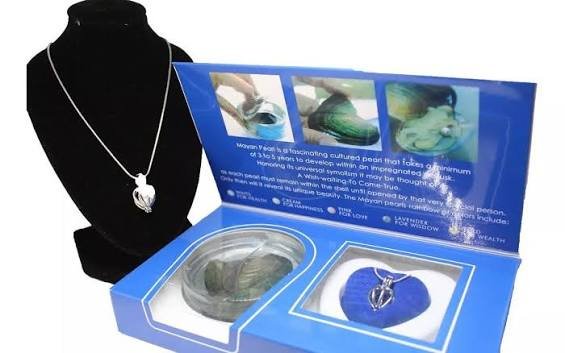 Estuche Mayan Pearl abierto con la ostra, el dije en forma de corazón y el libro informativo